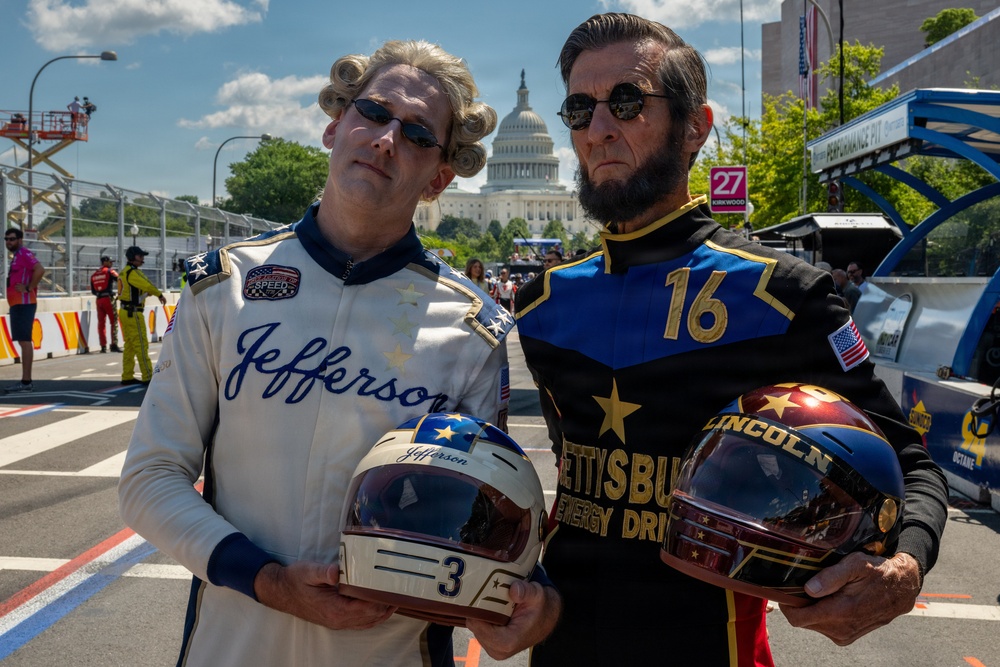 Two individuals dressed as historical U.S. presidents — Thomas Jefferson and Abraham Lincoln — are posing in racing suits at what appears to be a street racing event in Washington D.C., with the U.S. Capitol building visible in the background. Jefferson wears a white suit with number 3 and holds a matching helmet, while Lincoln wears a dark blue suit with number 16 sponsored by 'Gettysburg Energy Drink' and holds a gold-tinted helmet. The scene appears to be a promotional or entertainment appearance at a motorsport event.