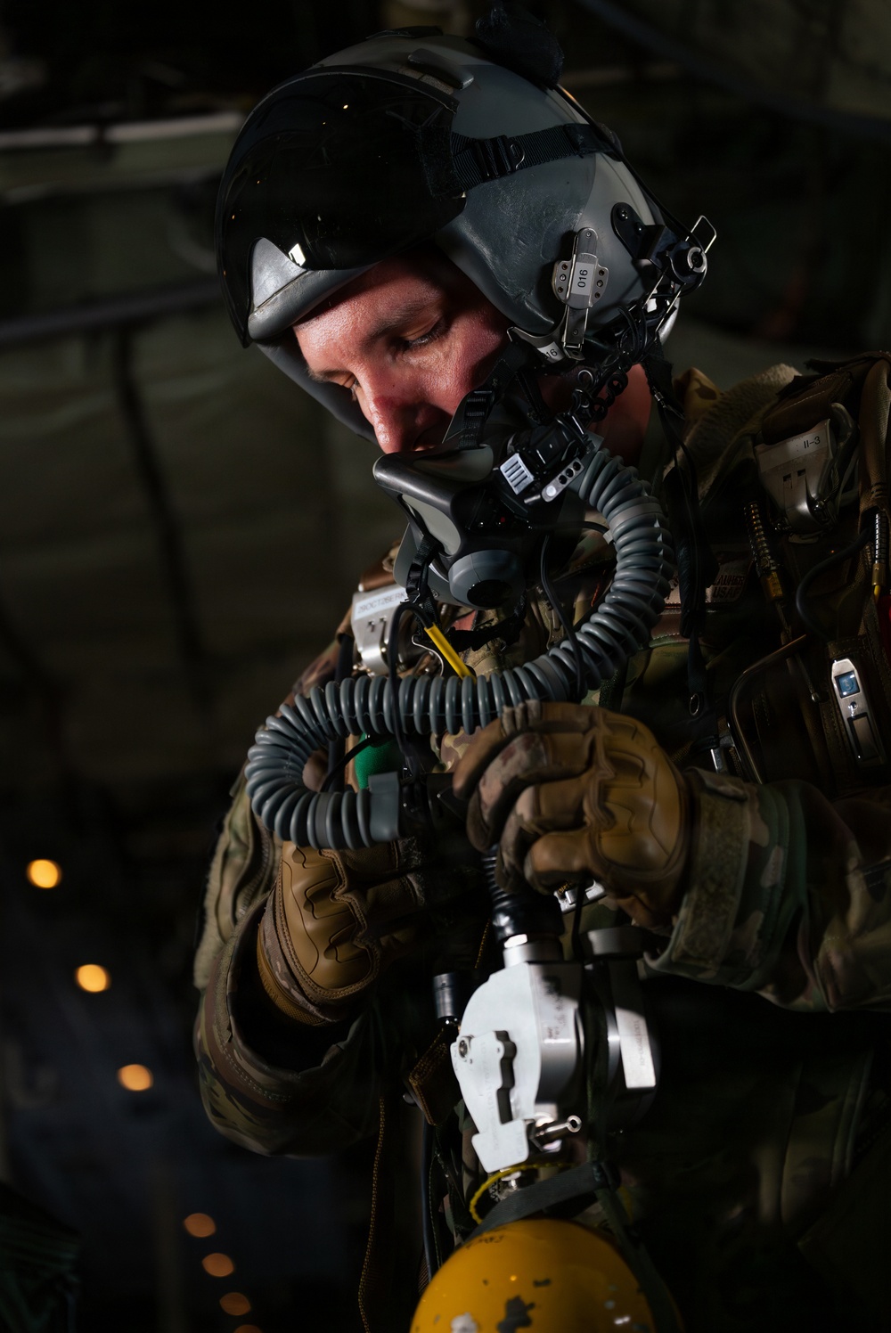 A military aviator or special operations personnel is shown in close-up, wearing full flight gear including a helmet with visor, oxygen mask with corrugated hose, and tactical vest. The individual appears to be checking or connecting oxygen/breathing equipment, possibly preparing for a high-altitude jump or flight operation. The dark background suggests the interior of a military aircraft.