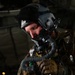 A military aviator or special operations personnel is shown in close-up, wearing full flight gear including a helmet with visor, oxygen mask with corrugated hose, and tactical vest. The individual appears to be checking or connecting oxygen/breathing equipment, possibly preparing for a high-altitude jump or flight operation. The dark background suggests the interior of a military aircraft.