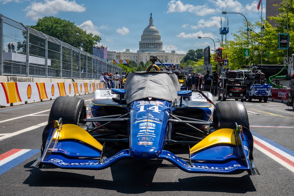 An IndyCar racing car numbered 14 is positioned in the pit lane or starting grid area with the United States Capitol building prominently visible in the background. The car is blue and gold with multiple sponsor logos and has a protective cover draped over the cockpit area. This appears to be the IndyCar Grand Prix on the Streets of Washington D.C., a street circuit race event.