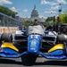 An IndyCar racing car numbered 14 is positioned in the pit lane or starting grid area with the United States Capitol building prominently visible in the background. The car is blue and gold with multiple sponsor logos and has a protective cover draped over the cockpit area. This appears to be the IndyCar Grand Prix on the Streets of Washington D.C., a street circuit race event.