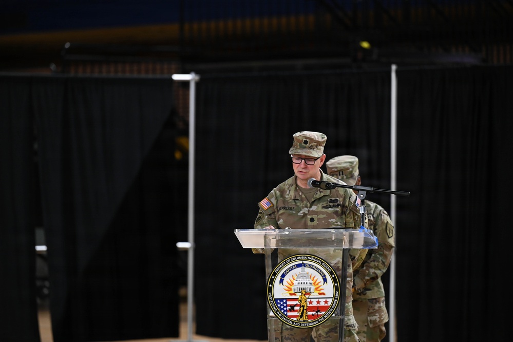 A U.S. Army officer in OCP (Operational Camouflage Pattern) uniform is speaking at a podium bearing the seal of the District of Columbia National Guard. The speaker appears to hold the rank of Lieutenant Colonel, indicated by the oak leaf insignia visible on the uniform. A second uniformed soldier stands in the background.