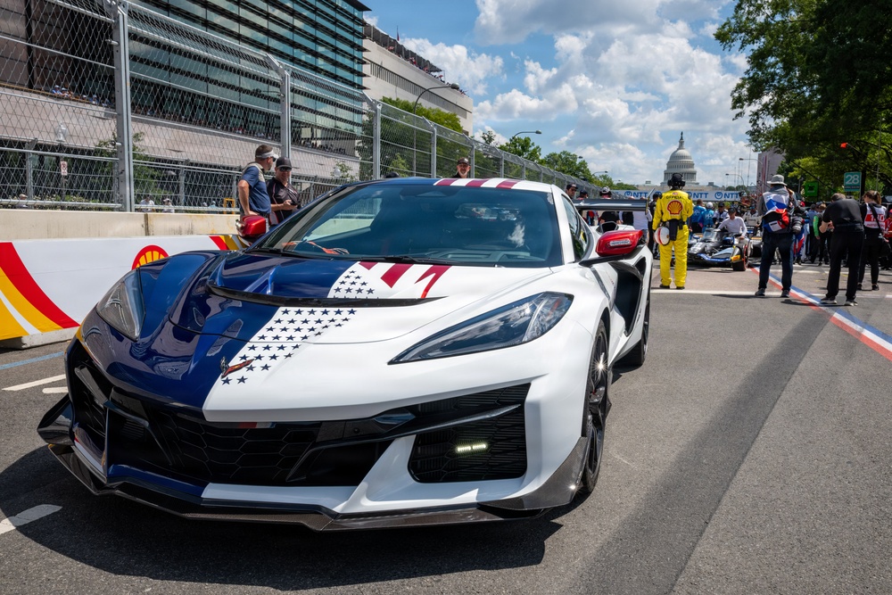 A Chevrolet Corvette Z06 with a patriotic American flag livery (red, white, and blue with stars and stripes) is positioned on a street circuit racing venue. The U.S. Capitol building is visible in the background, identifying the location as Washington D.C., consistent with the IndyCar Grand Prix on the Streets of Washington D.C. Race personnel, crew members, and IndyCar racing vehicles are visible in the background.
