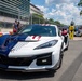 A Chevrolet Corvette Z06 with a patriotic American flag livery (red, white, and blue with stars and stripes) is positioned on a street circuit racing venue. The U.S. Capitol building is visible in the background, identifying the location as Washington D.C., consistent with the IndyCar Grand Prix on the Streets of Washington D.C. Race personnel, crew members, and IndyCar racing vehicles are visible in the background.
