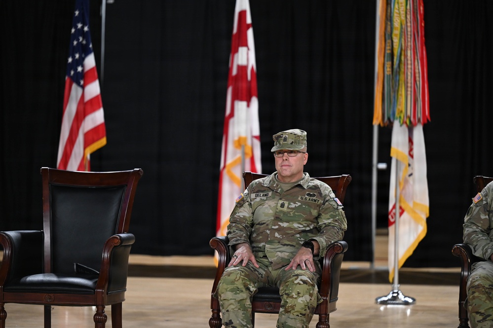 A U.S. Army soldier identified by name tape as 'DELAND' is seated in a formal wooden chair during what appears to be a military ceremony. He is wearing the Operational Camouflage Pattern (OCP) uniform with a senior NCO rank insignia visible on his cap. Multiple military flags and an American flag are displayed in the background, suggesting a change of command or similar formal military event.