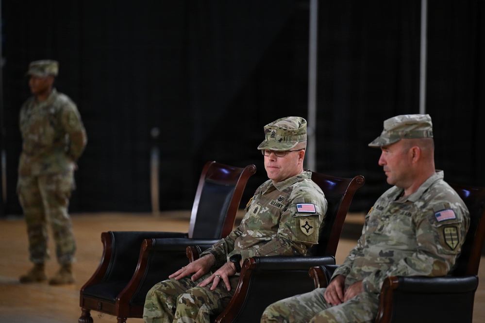 Three U.S. Army soldiers are present in what appears to be a formal military ceremony or event held indoors. Two soldiers are seated in ornate wooden chairs in the foreground, while a third stands in the background. All are wearing Army Combat Uniforms (ACU) in MultiCam/OCP pattern with U.S. flag patches and unit insignia visible.