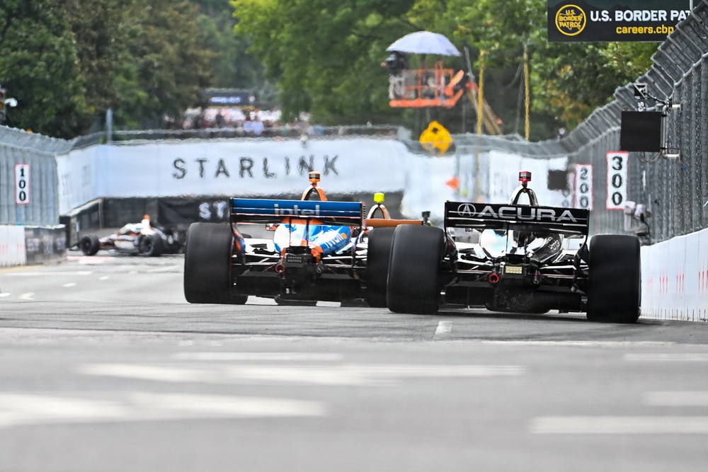 Two IndyCar racing cars are competing side by side on a street circuit, viewed from a rear angle. The cars are approaching a braking zone marked with distance boards (300, 200, 100 meters). The race takes place on a temporary street circuit with concrete barriers, featuring sponsor signage including Starlink and U.S. Border Patrol.