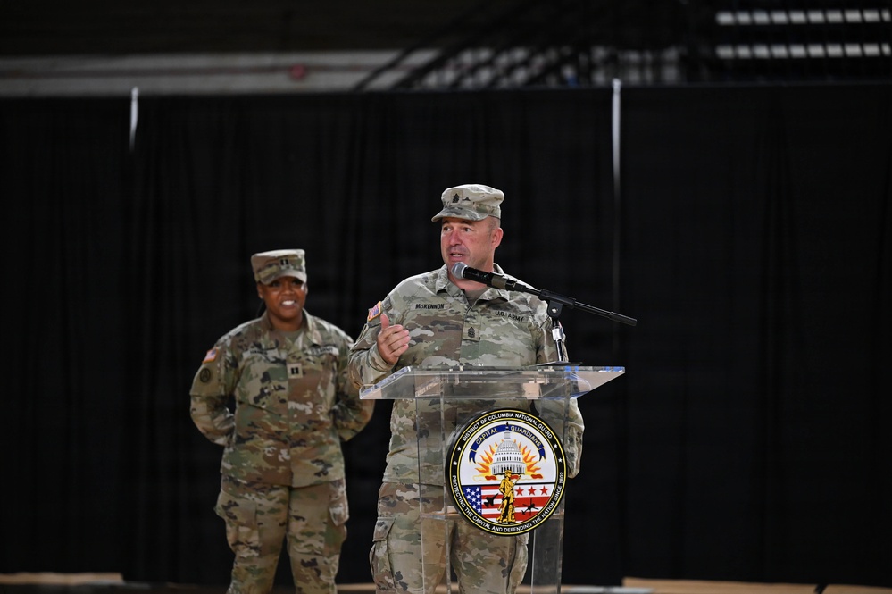 Two U.S. Army soldiers in OCP (Operational Camouflage Pattern) uniforms are present at an official military event. The male soldier at the podium, identified by his name tape as 'McKENNON,' is delivering a speech at a District of Columbia National Guard event. A female soldier wearing captain's rank insignia stands in the background.