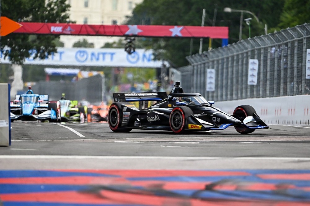 An IndyCar race is taking place on a street circuit, with a black car prominently in the foreground displaying 'OpenAI' sponsorship. Multiple race cars are visible in the background on what appears to be a temporary street course. Sponsor banners including 'NTT' and 'FOX' are visible on overhead gantries.