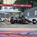 An IndyCar race is taking place on a street circuit, with a black car prominently in the foreground displaying 'OpenAI' sponsorship. Multiple race cars are visible in the background on what appears to be a temporary street course. Sponsor banners including 'NTT' and 'FOX' are visible on overhead gantries.