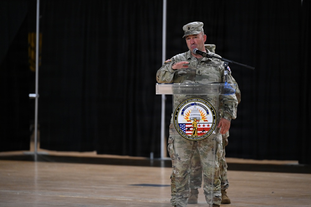 A U.S. Army soldier in OCP (Operational Camouflage Pattern) uniform is speaking at a podium bearing the seal of the District of Columbia National Guard. The setting appears to be an indoor military ceremony or briefing held in a large hall or armory. A second uniformed individual is partially visible in the background.