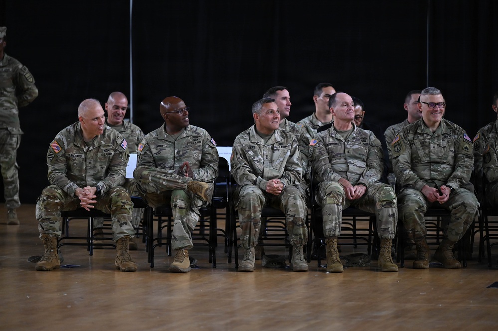 A group of U.S. Army soldiers in OCP (Operational Camouflage Pattern) uniforms are seated in chairs, appearing to be attending a formal military ceremony or event. The front row consists of approximately five senior soldiers, several of whom are smiling and appear engaged with something off-camera. A second row of soldiers is visible behind them in a darkened indoor venue with a wooden floor.
