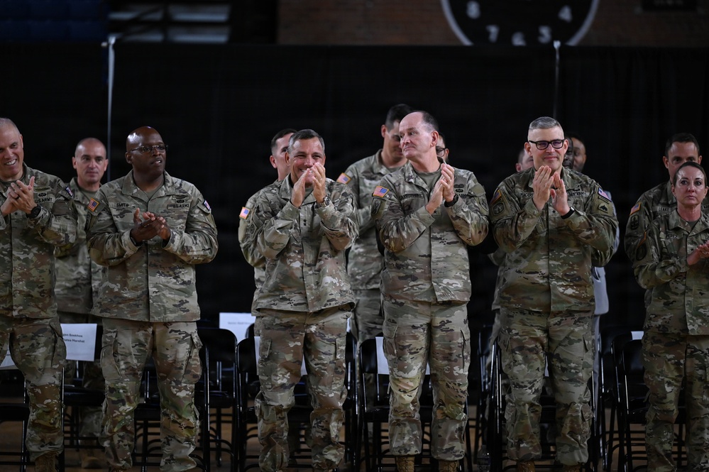 A group of U.S. Army soldiers in Operational Camouflage Pattern (OCP) uniforms are standing and applauding at what appears to be a formal military ceremony or event. The soldiers appear to be senior-ranking personnel based on their insignia and positioning. The setting appears to be an indoor venue or hangar with dim background lighting.