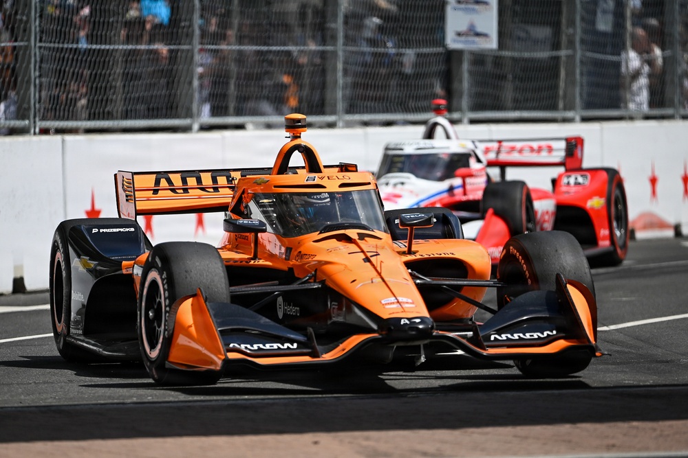 An IndyCar race is underway on a street circuit, with a prominent orange Arrow McLaren car leading in the foreground. A red and white car follows closely behind in the background. Spectators are visible behind safety fencing along the track barrier.