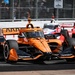 An IndyCar race is underway on a street circuit, with a prominent orange Arrow McLaren car leading in the foreground. A red and white car follows closely behind in the background. Spectators are visible behind safety fencing along the track barrier.
