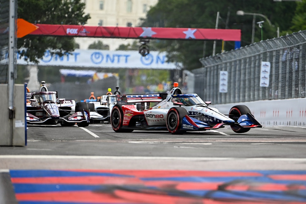 An IndyCar racing event taking place on a street circuit, with multiple open-wheel race cars navigating a straightaway. The foreground features a red, white, and blue livery car with 'Orion180 Insurance' and 'REcreative' sponsorship markings. Sponsor banners including NTT and FOX are visible overhead, along with a patriotic star-themed gantry banner.