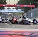 An IndyCar racing event taking place on a street circuit, with multiple open-wheel race cars navigating a straightaway. The foreground features a red, white, and blue livery car with 'Orion180 Insurance' and 'REcreative' sponsorship markings. Sponsor banners including NTT and FOX are visible overhead, along with a patriotic star-themed gantry banner.