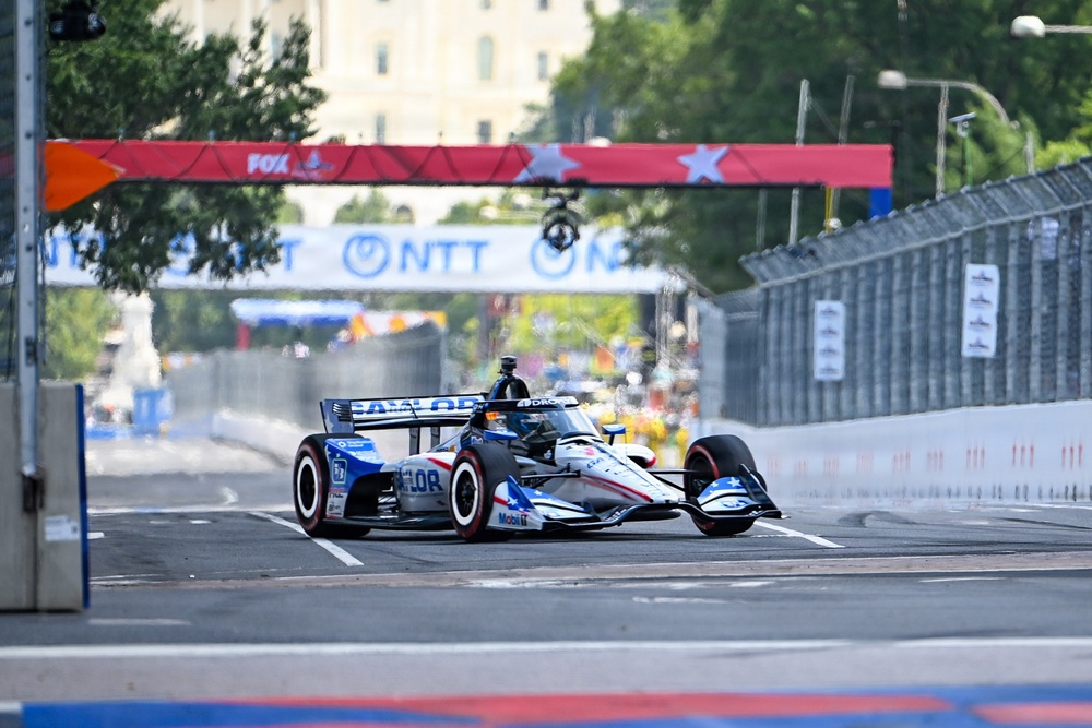 An IndyCar racing vehicle navigates a street circuit course, identifiable by the temporary barriers and sponsor banners lining the track. The car features a blue, white, and red livery with visible sponsorships including 'Mobil 1' and 'Taylor' branding. Overhead signage includes NTT and FOX branding, suggesting this is an NTT IndyCar Series event on a temporary street circuit.