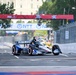 An IndyCar racing vehicle navigates a street circuit course, identifiable by the temporary barriers and sponsor banners lining the track. The car features a blue, white, and red livery with visible sponsorships including 'Mobil 1' and 'Taylor' branding. Overhead signage includes NTT and FOX branding, suggesting this is an NTT IndyCar Series event on a temporary street circuit.