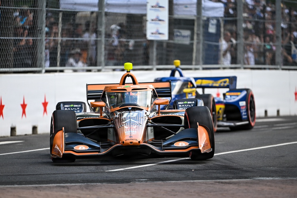 Two IndyCar open-wheel racing cars are competing on a street circuit, with the foreground car featuring a distinctive orange and black livery with Bitcoin and 'Only Bulls' sponsorship branding. The race appears to be taking place in Chicago, as evidenced by the red star motifs on the barrier walls, consistent with the Chicago Street Race. Spectators are visible behind safety fencing in the background.
