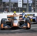 Two IndyCar open-wheel racing cars are competing on a street circuit, with the foreground car featuring a distinctive orange and black livery with Bitcoin and 'Only Bulls' sponsorship branding. The race appears to be taking place in Chicago, as evidenced by the red star motifs on the barrier walls, consistent with the Chicago Street Race. Spectators are visible behind safety fencing in the background.