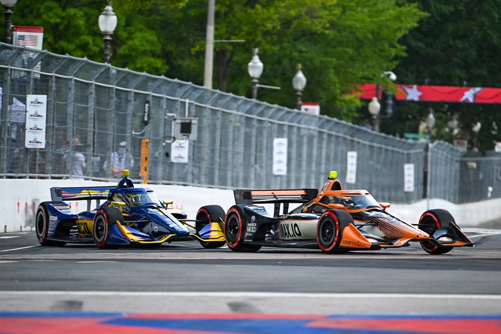 Three IndyCar open-wheel racing cars are competing side by side on a street circuit, with temporary metal barriers lining the course. The cars display distinct liveries: blue/yellow, black/silver, and orange. The setting appears to be an urban street race venue with lamp posts, trees, and spectator infrastructure visible in the background.