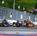 Three IndyCar open-wheel racing cars are competing side by side on a street circuit, with temporary metal barriers lining the course. The cars display distinct liveries: blue/yellow, black/silver, and orange. The setting appears to be an urban street race venue with lamp posts, trees, and spectator infrastructure visible in the background.