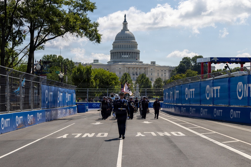 A military marching band or honor guard is proceeding down a temporary racing circuit street course in Washington D.C., with the United States Capitol Building prominently visible in the background. The track is lined with blue NTT-branded barriers on both sides, indicating this is an IndyCar or similar motorsport event. The scene appears to be a pre-race ceremonial procession.