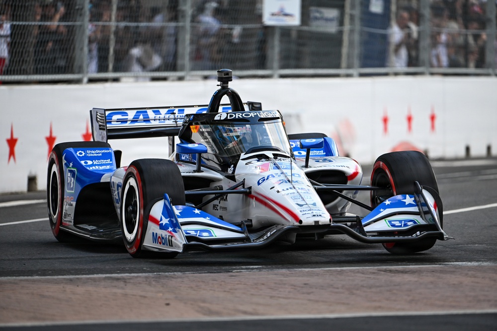An IndyCar racing vehicle with a patriotic red, white, and blue livery featuring stars and stripes is captured navigating a street circuit. The car displays multiple sponsor logos including Mobil 1, ShipStation Global, United Rentals, Droplight, and Gayle/Gaylor branding. Spectators and safety fencing are visible in the background, indicating an active race event.
