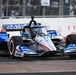 An IndyCar racing vehicle with a patriotic red, white, and blue livery featuring stars and stripes is captured navigating a street circuit. The car displays multiple sponsor logos including Mobil 1, ShipStation Global, United Rentals, Droplight, and Gayle/Gaylor branding. Spectators and safety fencing are visible in the background, indicating an active race event.