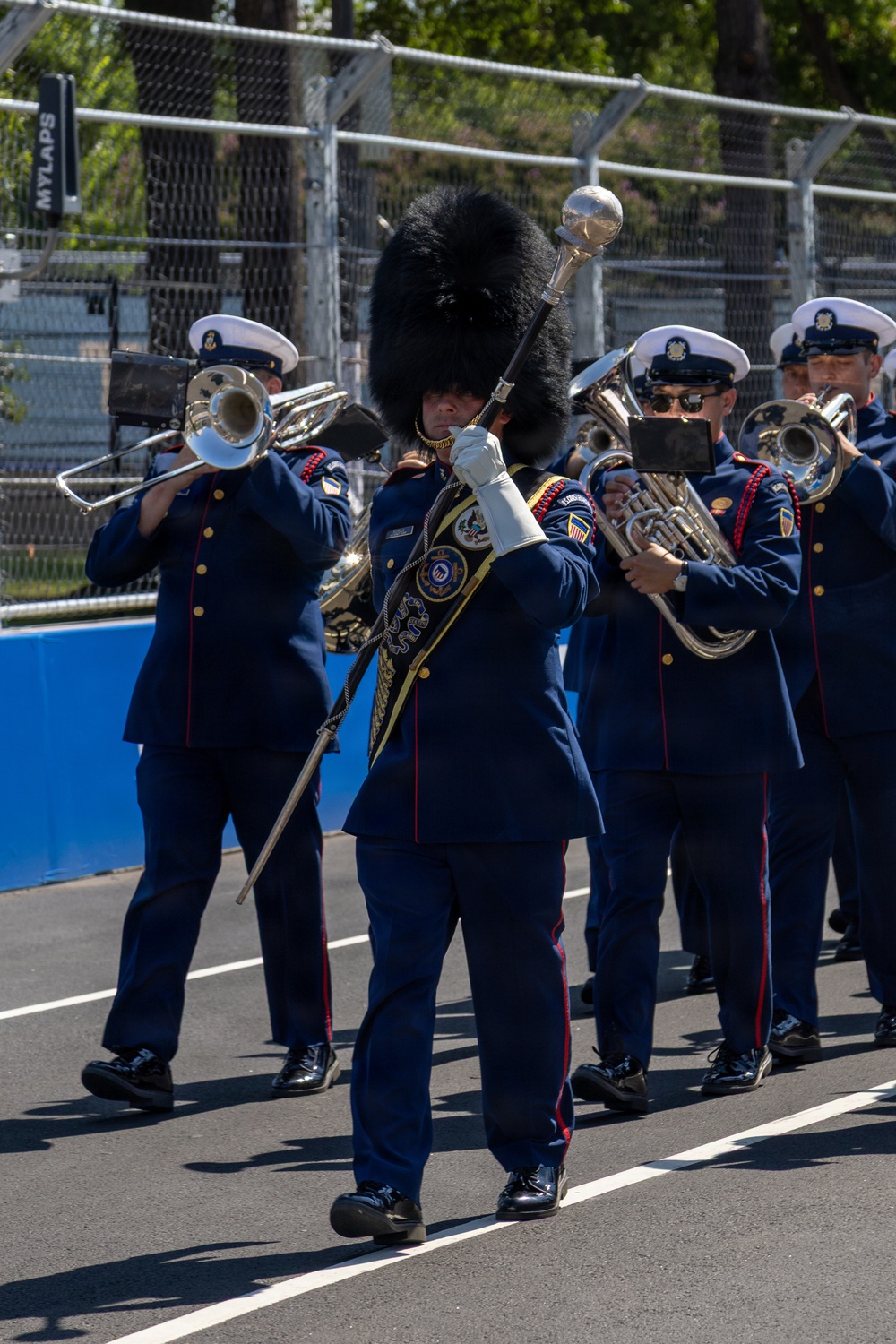A military marching band, identifiable as the United States Coast Guard Band, is performing on what appears to be a racing circuit or street course. The drum major leads the procession wearing a distinctive tall black bearskin hat and carrying a ceremonial mace, while band members in navy blue dress uniforms with red trim play brass instruments. A 'MYLAPS' timing system sign is visible in the background, confirming a motorsport venue setting.