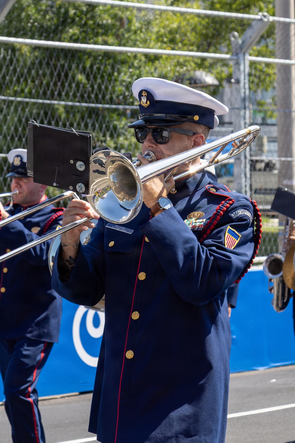 A U.S. Coast Guard band member plays a trombone during what appears to be an outdoor parade or ceremonial performance. The musician is dressed in full dress uniform with a white-topped service cap, navy blue coat with red trim, gold buttons, service ribbons, and a U.S. Coast Guard patch on the sleeve. A second band member is partially visible in the background.