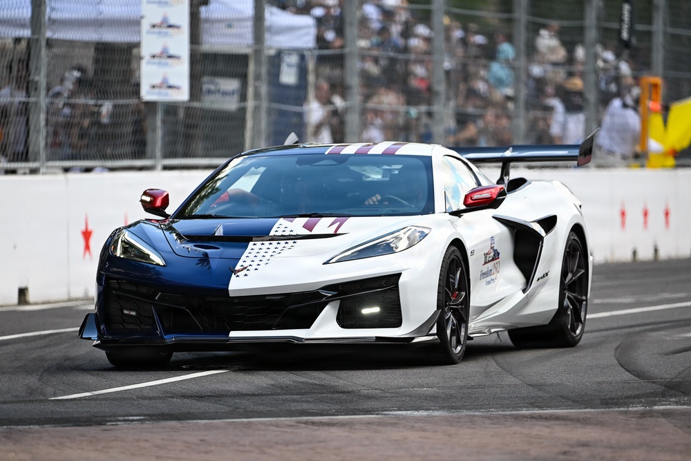 A Chevrolet Corvette Z06 with a patriotic red, white, and blue livery is driving on a racing circuit. The car features a split blue and white color scheme with star motifs and purple racing stripes, along with a large rear wing. Spectators and safety barriers are visible in the background, suggesting a professional racing or pace car event.