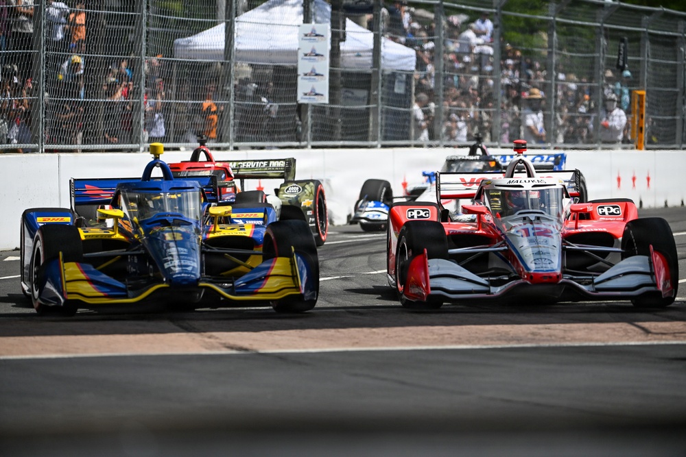An IndyCar racing event captured mid-race showing multiple open-wheel cars competing side by side on a street circuit. The foreground features two prominent cars — one in blue and yellow livery with DHL sponsorship, and one in red, white, and silver with PPG sponsorship. Spectators line the fenced barriers in the background, and additional race cars are visible further down the track.
