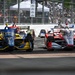 An IndyCar racing event captured mid-race showing multiple open-wheel cars competing side by side on a street circuit. The foreground features two prominent cars — one in blue and yellow livery with DHL sponsorship, and one in red, white, and silver with PPG sponsorship. Spectators line the fenced barriers in the background, and additional race cars are visible further down the track.