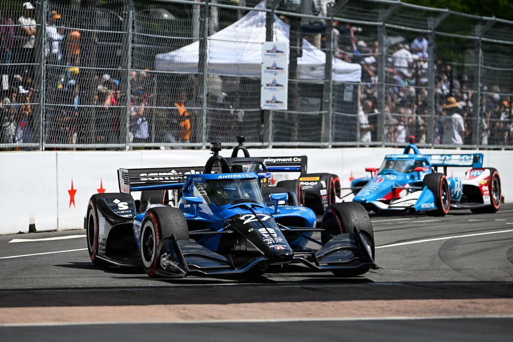 An IndyCar racing event captured on a street circuit, showing multiple open-wheel race cars navigating a turn. The foreground features car number 27 from Andretti Autosport in a blue and black livery with VIZIO and Sonsio sponsorship. A large crowd of spectators is visible behind safety fencing in the background.