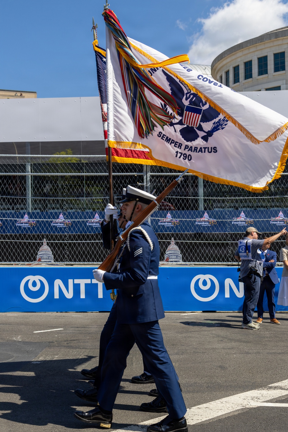 A military color guard is marching in a ceremonial procession at what appears to be the 'Freedom 250 Grand Prix' racing event. The guard carries multiple flags including a United States Coast Guard flag bearing the motto 'Semper Paratus 1790' and the American flag. The event is sponsored by NTT and features race track barriers and fencing in the background.