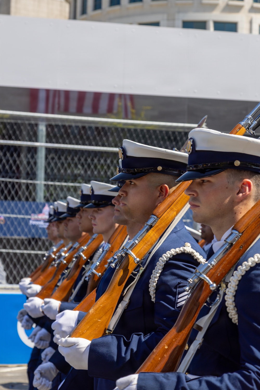 A military honor guard unit stands in precise formation during a ceremonial event. The personnel are dressed in formal navy blue dress uniforms with white-topped caps and white gloves, each carrying a rifle with a wooden stock and chrome fittings. The image captures the disciplined alignment of the formation from a side-profile perspective.