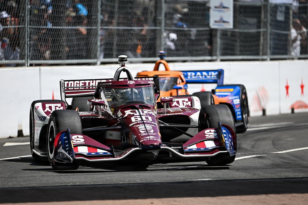 An IndyCar race is underway on a street circuit, with car number 60 prominently featured in the foreground. The lead car is decorated in a dark maroon/crimson livery with Texas A&amp;M (ATM) university branding and 'TOGETHER' text on the rear wing. At least two other IndyCar competitors are visible behind it, including an orange McLaren-liveried car and a blue US Bank-sponsored car.