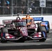 An IndyCar race is underway on a street circuit, with car number 60 prominently featured in the foreground. The lead car is decorated in a dark maroon/crimson livery with Texas A&amp;M (ATM) university branding and 'TOGETHER' text on the rear wing. At least two other IndyCar competitors are visible behind it, including an orange McLaren-liveried car and a blue US Bank-sponsored car.