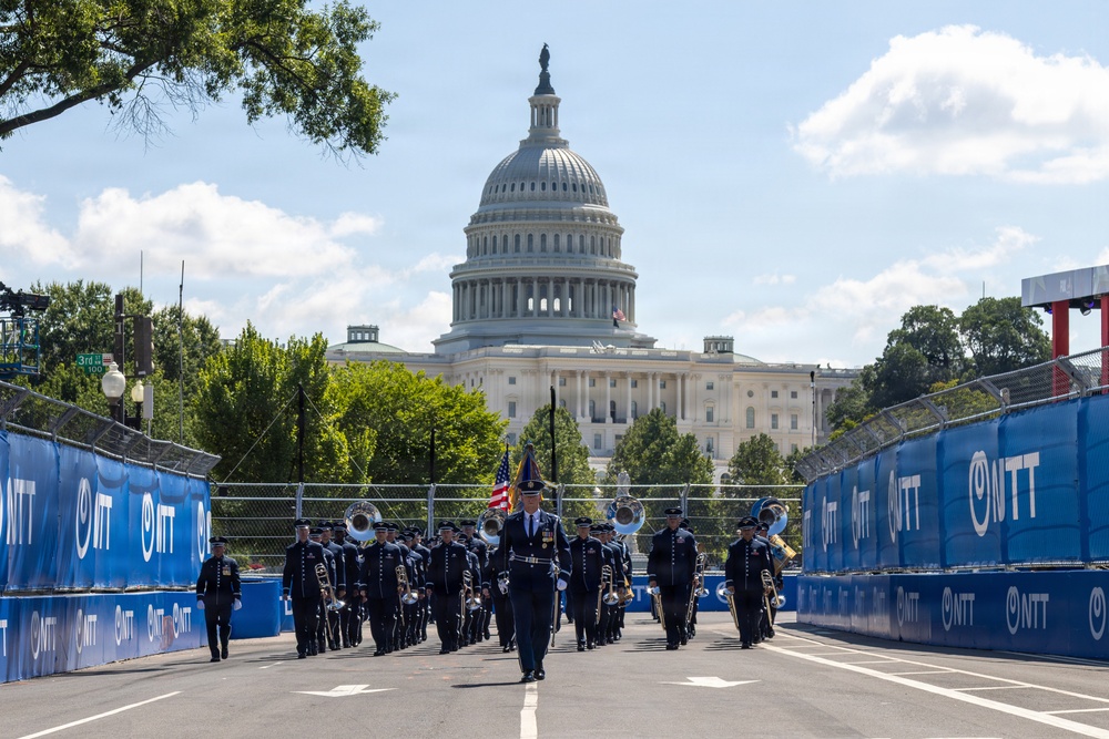 A military marching band parades down a closed street in Washington D.C., with the United States Capitol Building prominently visible in the background. The street is lined with blue NTT-branded barriers, suggesting this is part of a motorsport or major public event setup. The procession is led by a uniformed officer carrying an American flag.