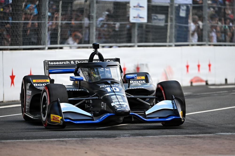 An IndyCar racing car numbered 10 is captured mid-race on a street circuit. The black and blue livery car features sponsorships from OpenAI, Root Insurance, DHL, and 'Research &amp; Donkerman Co.' Spectators and safety barriers are visible in the background.