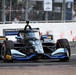 An IndyCar racing car numbered 10 is captured mid-race on a street circuit. The black and blue livery car features sponsorships from OpenAI, Root Insurance, DHL, and 'Research &amp; Donkerman Co.' Spectators and safety barriers are visible in the background.