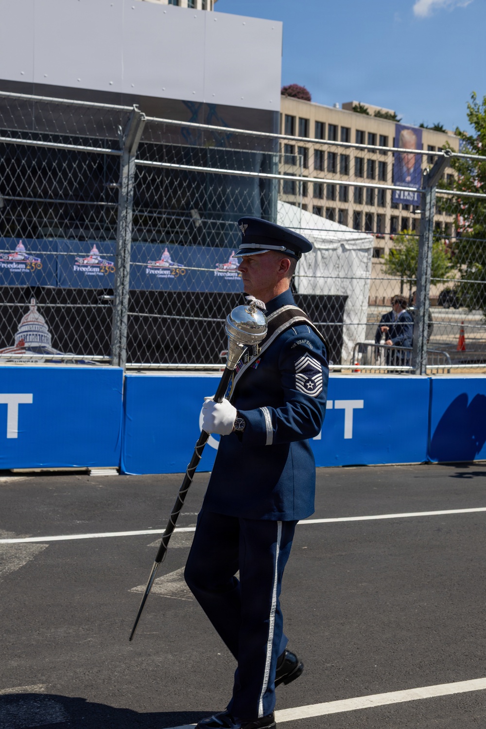 A U.S. Air Force Band drum major in full ceremonial dress uniform marches while carrying a mace on what appears to be a street course. The event is the 'Freedom 250 Grand Prix' based on visible signage, set in an urban environment with security fencing and grandstand infrastructure. The U.S. Capitol building is visible on event branding in the background.