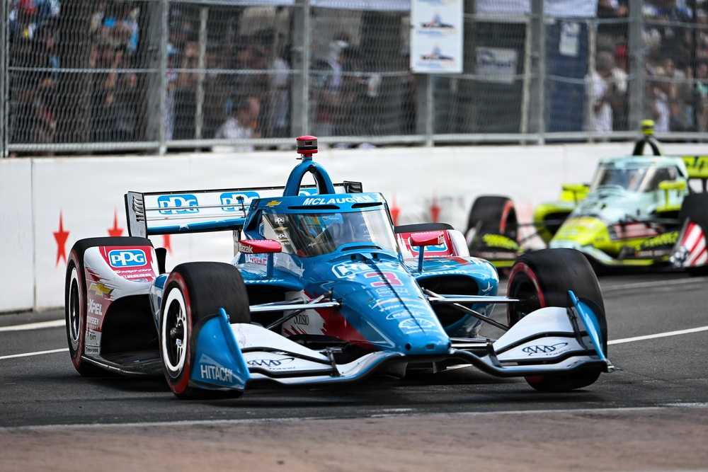An IndyCar race is underway on a street circuit, with a blue and white PPG/Hitachi-sponsored car prominently in the foreground. The lead car bears 'McLaughlin' branding on the halo and is sponsored by PPG and Hitachi. A second yellow-green car is visible in the background, with spectators lining the fenced barriers.