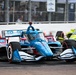 An IndyCar race is underway on a street circuit, with a blue and white PPG/Hitachi-sponsored car prominently in the foreground. The lead car bears 'McLaughlin' branding on the halo and is sponsored by PPG and Hitachi. A second yellow-green car is visible in the background, with spectators lining the fenced barriers.