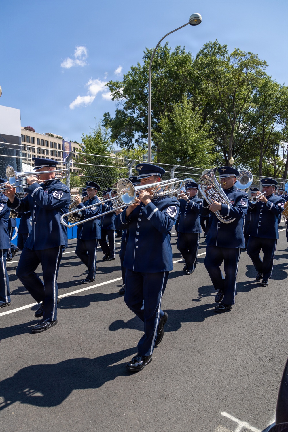 A military marching band, identifiable as United States Air Force personnel based on their distinctive blue dress uniforms with silver trim and Air Force insignia caps, performs while marching down a paved street. The musicians are playing brass instruments including trombones, euphoniums, and trumpets in a formal parade formation. The setting appears to be an urban outdoor venue with chain-link security fencing, trees, and buildings visible in the background under a partly cloudy sky.