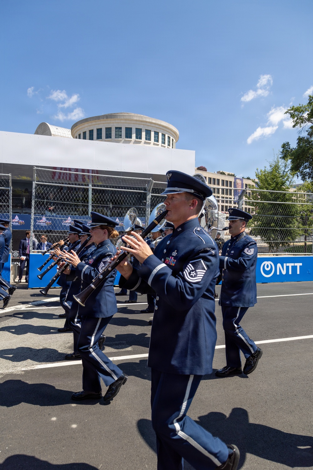 A U.S. Air Force marching band is performing in an outdoor parade or ceremonial event on a paved street. Band members are dressed in formal dark blue Air Force dress uniforms with white trim and are playing woodwind and brass instruments while marching. The event appears to take place near a venue with NTT sponsorship signage and chain-link fencing in the background.
