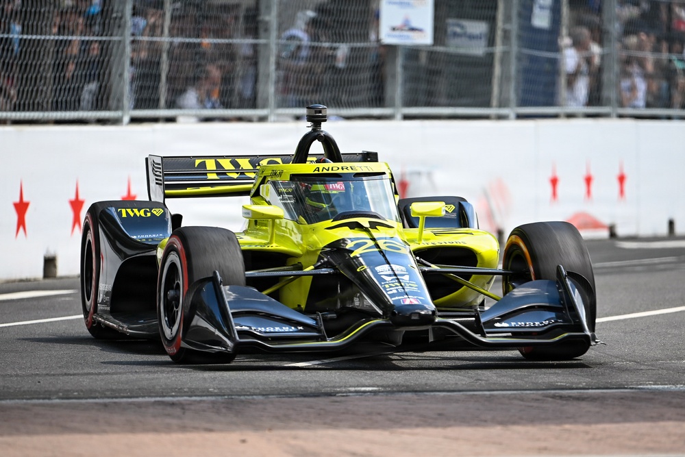 An IndyCar racing vehicle with a distinctive yellow-green and black livery is captured on a street circuit during a race or practice session. The car prominently displays 'TWG' and 'Andretti' branding, indicating it belongs to Andretti Autosport. Spectators and safety fencing are visible in the background.