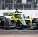 An IndyCar racing vehicle with a distinctive yellow-green and black livery is captured on a street circuit during a race or practice session. The car prominently displays 'TWG' and 'Andretti' branding, indicating it belongs to Andretti Autosport. Spectators and safety fencing are visible in the background.